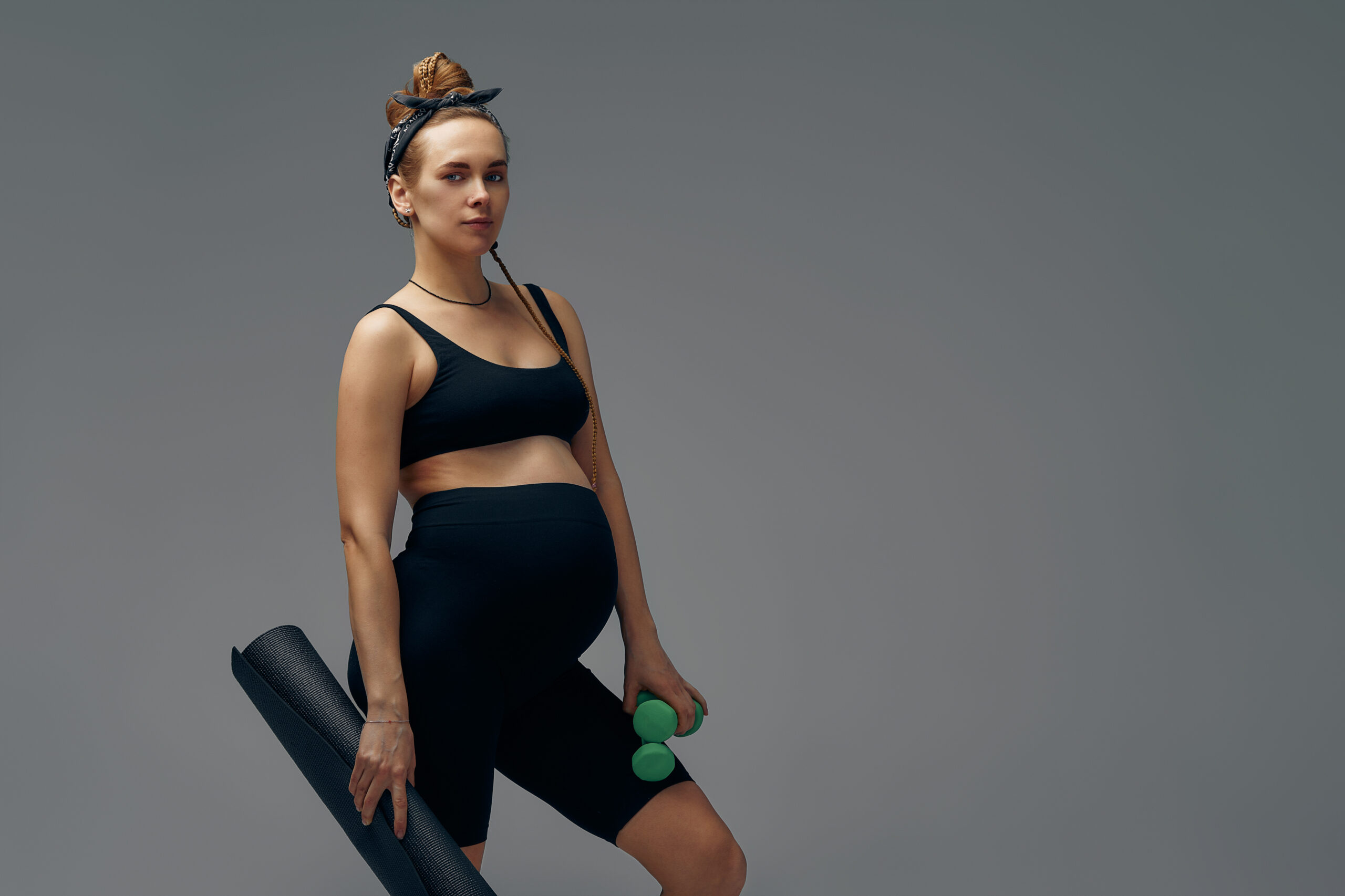 Fit young woman exercising during her pregnancy standing smiling at the camera with bottled water in one hand and a gym mat in the other.