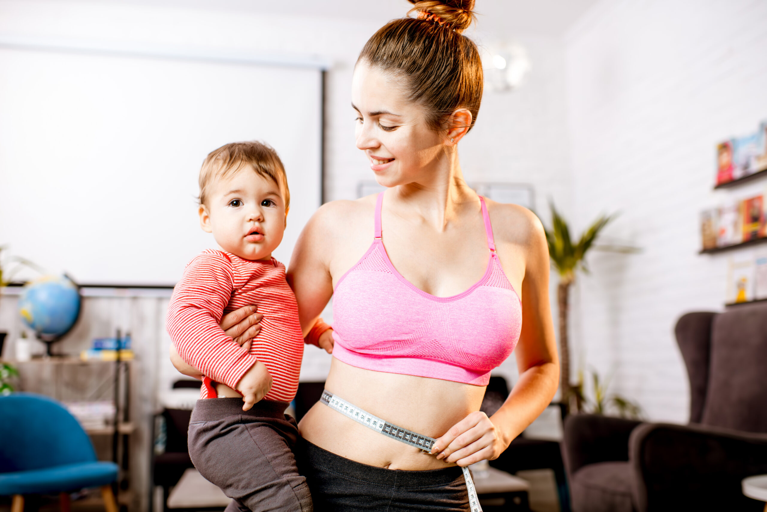 Young mother in sportswear measuring her waist worried about her weight after the child birth sitting with her baby son during the exercise