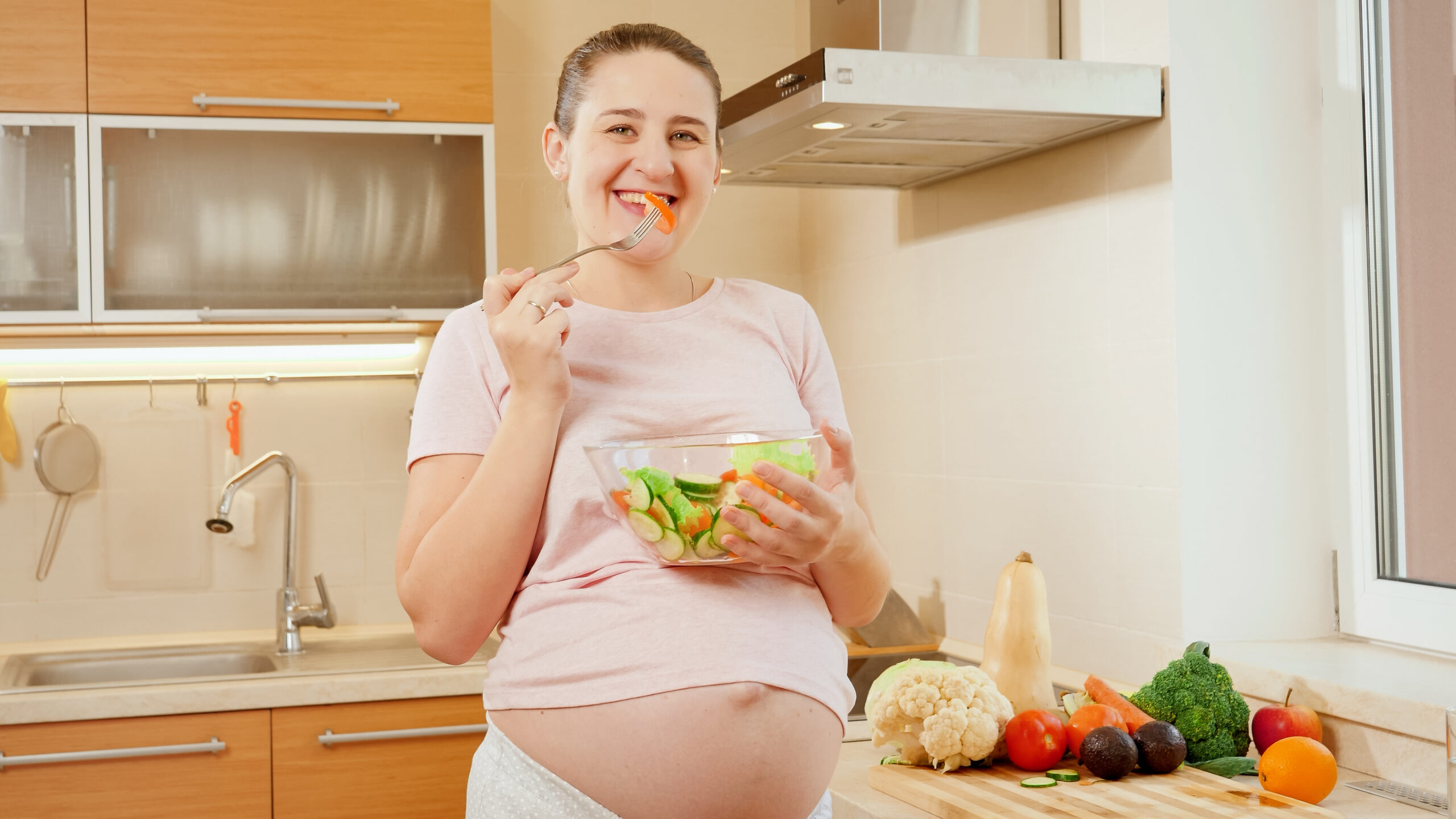 Portrait of smiling pregnant woman holding big glass bowl and eating vegetable salad on kitchen.