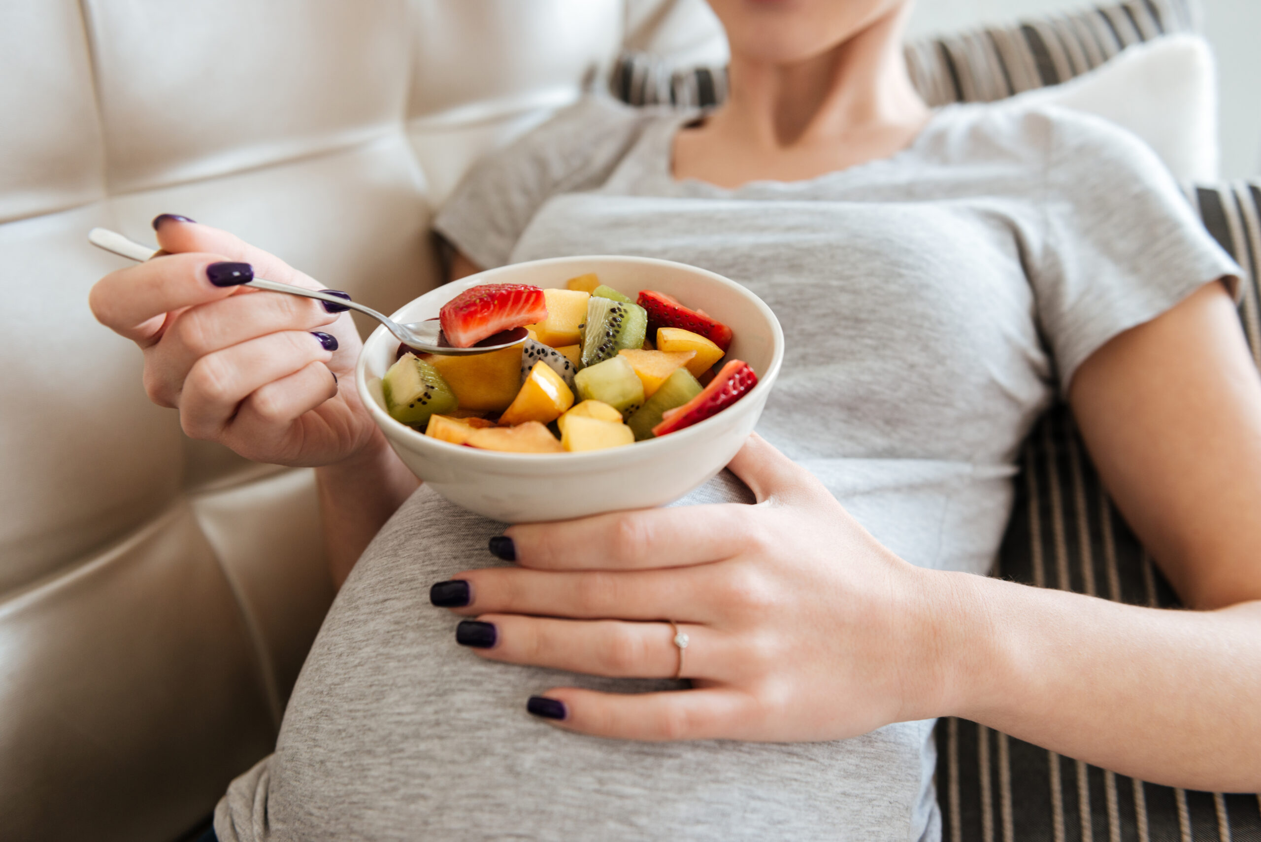 Closeup of pregnant young woman sitting on sofa and eating fruit salad at home
